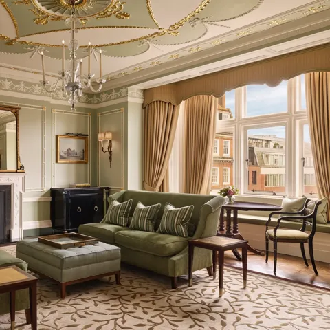 An elegant sitting room with sage green walls, gold detailing, and classic furnishings including velvet sofas, a marble fireplace, chandelier, and large window overlooking red-brick Mayfair buildings.