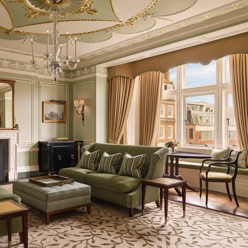 An elegant sitting room with sage green walls, gold detailing, and classic furnishings including velvet sofas, a marble fireplace, chandelier, and large window overlooking red-brick Mayfair buildings.