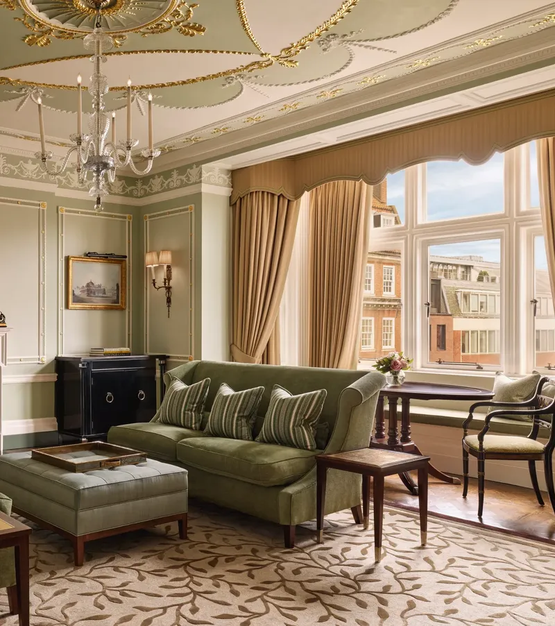 An elegant sitting room with sage green walls, gold detailing, and classic furnishings including velvet sofas, a marble fireplace, chandelier, and large window overlooking red-brick Mayfair buildings.