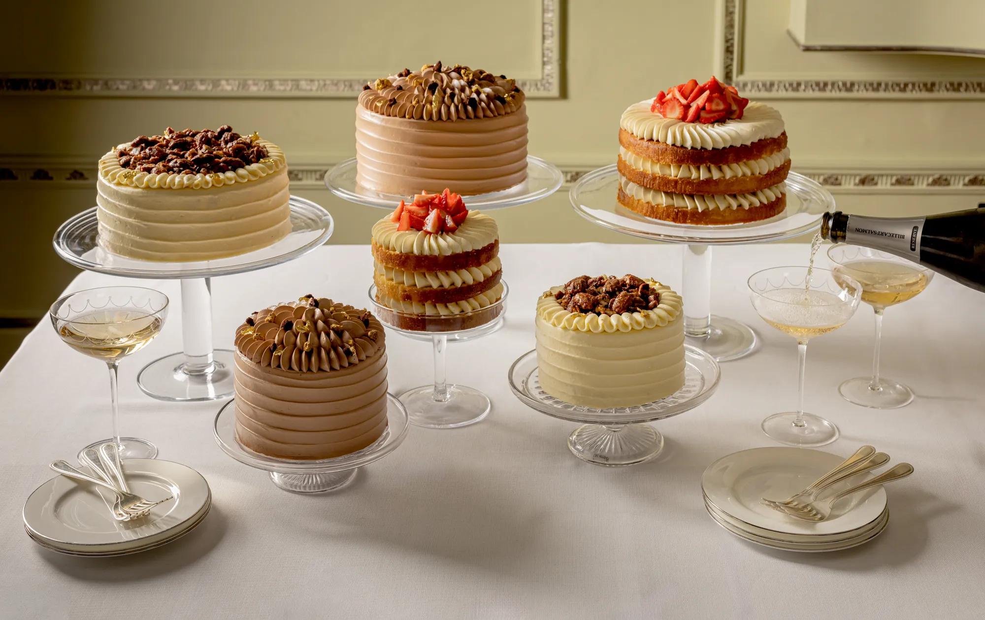 Five decorated celebration cakes displayed on glass stands around a table, with champagne being poured into a coupe glass.