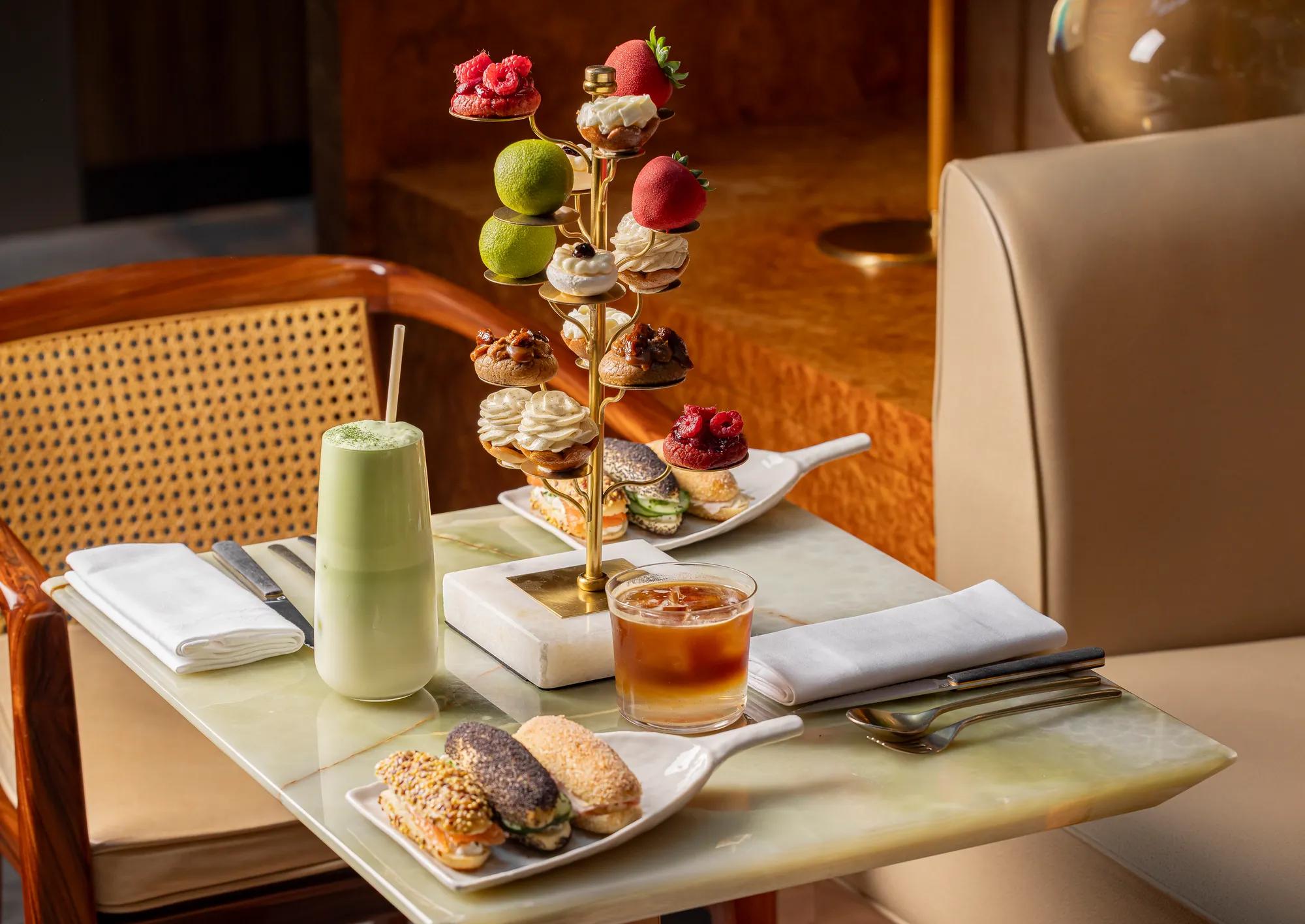 Gold pastry stand with fruit-shaped and piped cream pastries, alongside matcha latte, iced coffee, and eclairs on a marble table.