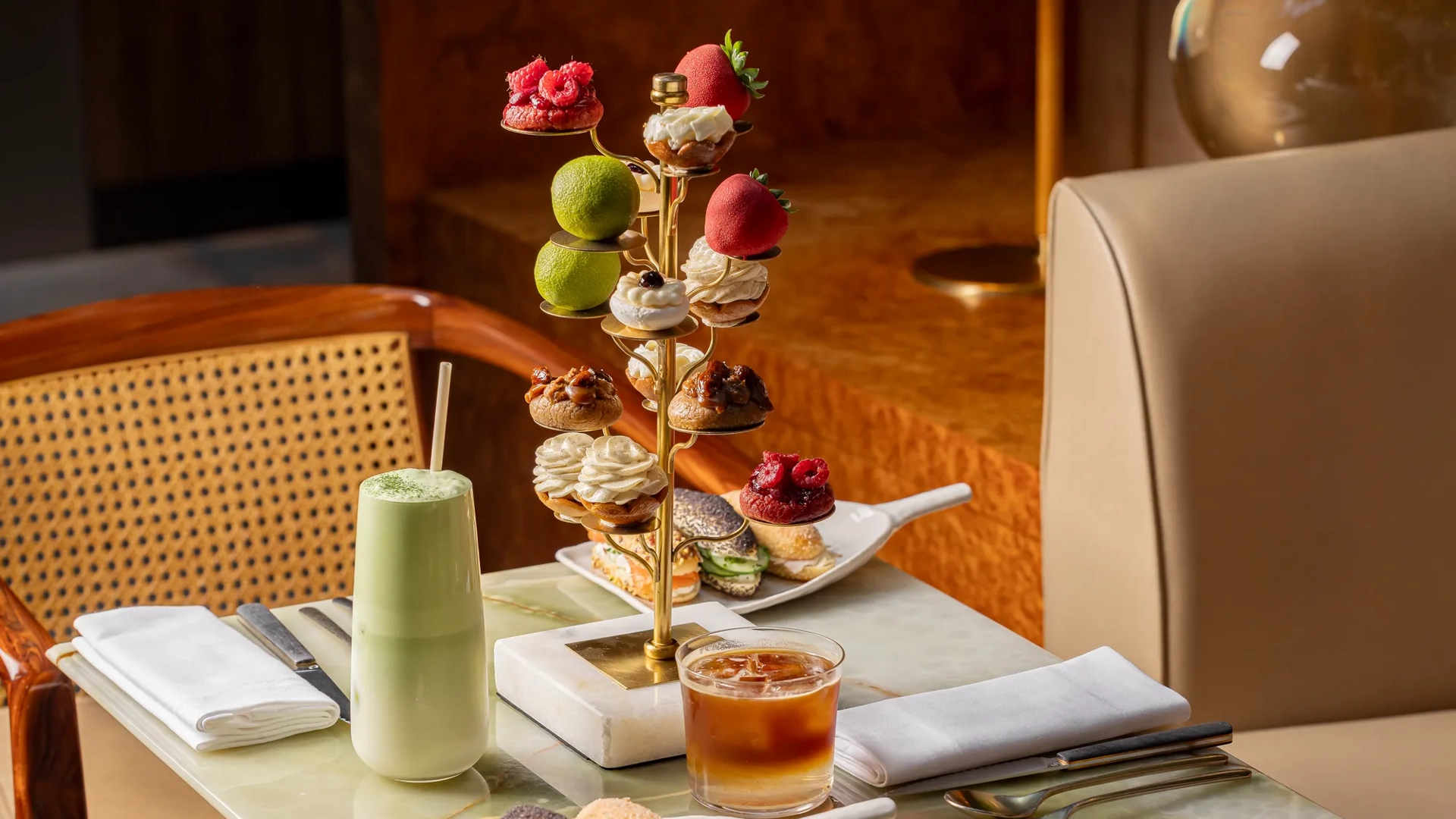 Gold pastry stand with fruit-shaped and piped cream pastries, alongside matcha latte, iced coffee, and eclairs on a marble table.