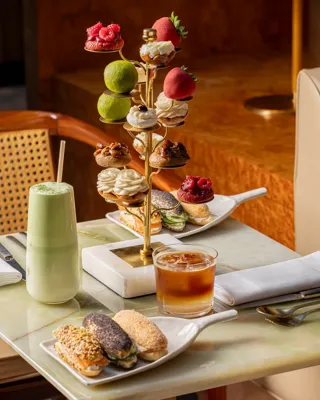 Gold pastry stand with fruit-shaped and piped cream pastries, alongside matcha latte, iced coffee, and eclairs on a marble table.