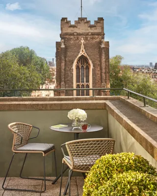 Terrace with round table, chairs, flowers, and city view featuring historic church tower.