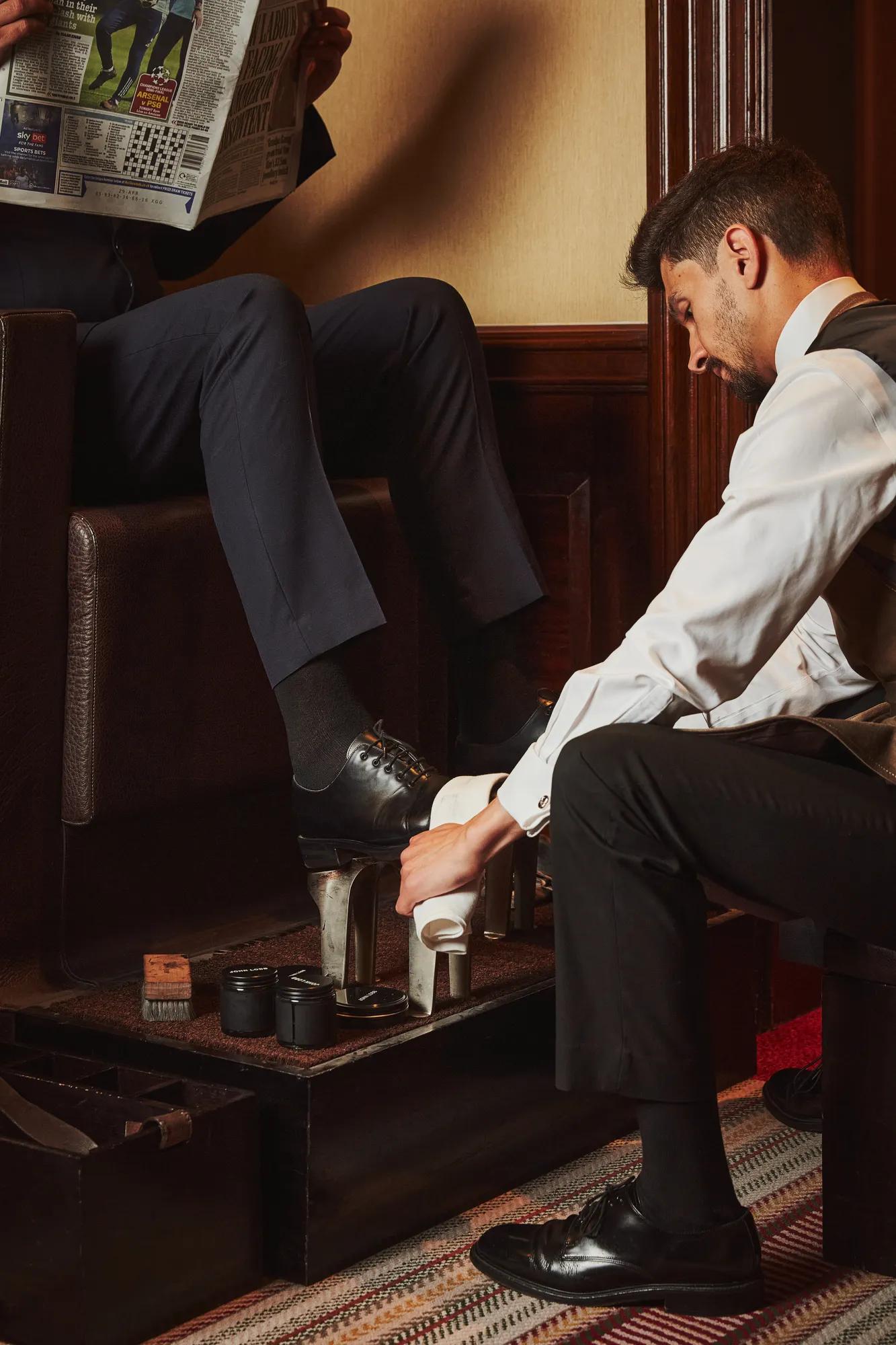 Valet polishing a guest’s black dress shoes at a traditional shoeshine stand as the guest reads a newspaper.