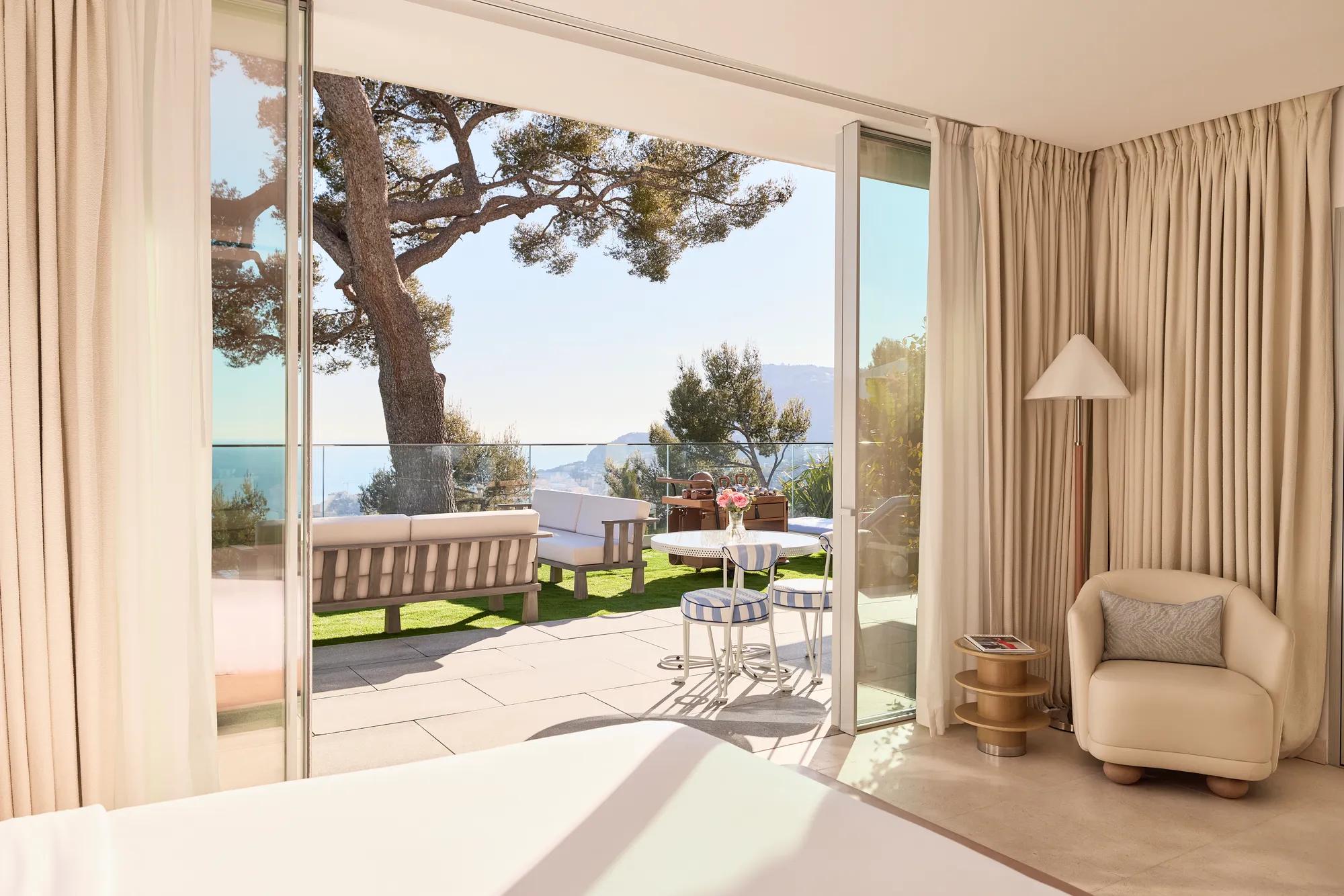 Suite bedroom with floor-to-ceiling glass doors opening to a private terrace. A mature pine tree frames views of the Mediterranean coastline and distant mountains.