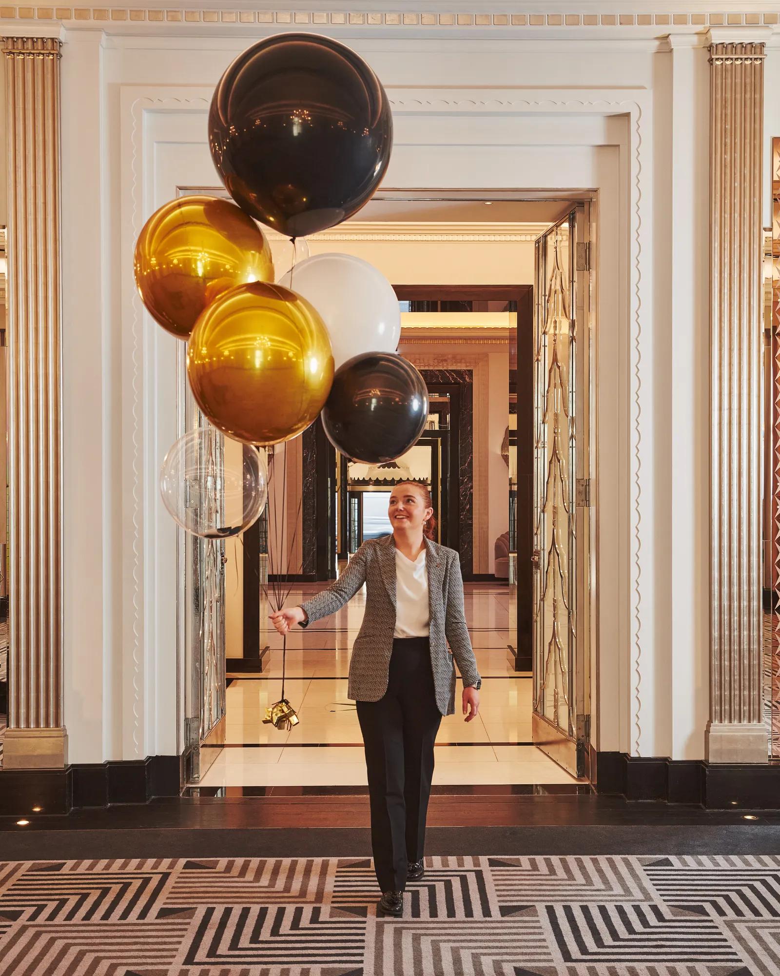A person walks through Claridge’s Art Deco hallway holding a cluster of gold, black, white, and clear balloons, surrounded by mirrored doors and geometric patterned flooring.