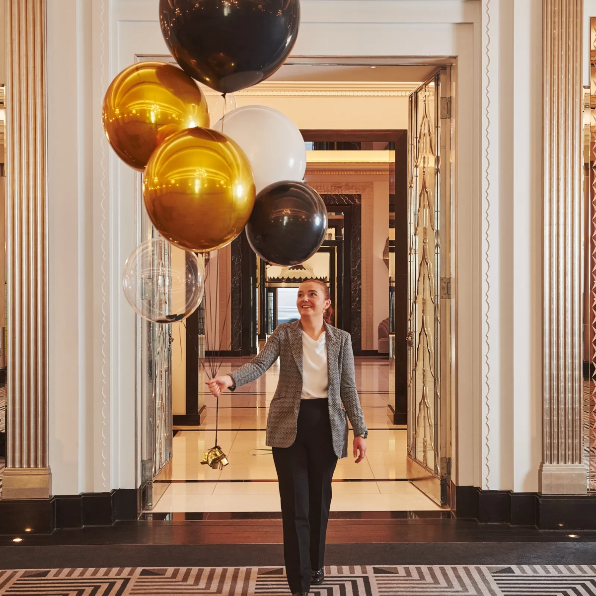 A person walks through Claridge’s Art Deco hallway holding a cluster of gold, black, white, and clear balloons, surrounded by mirrored doors and geometric patterned flooring.