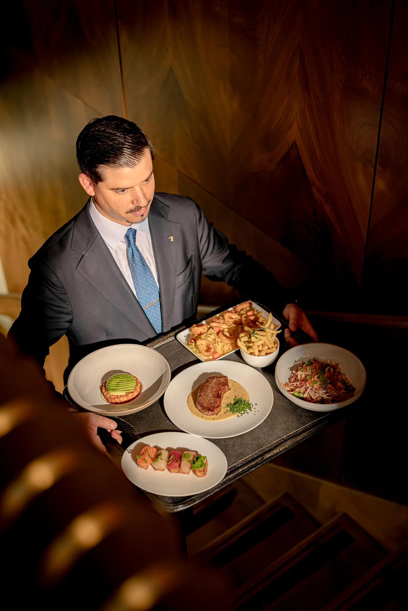 Staff member carrying a tray of restaurant dishes through a hotel interior.