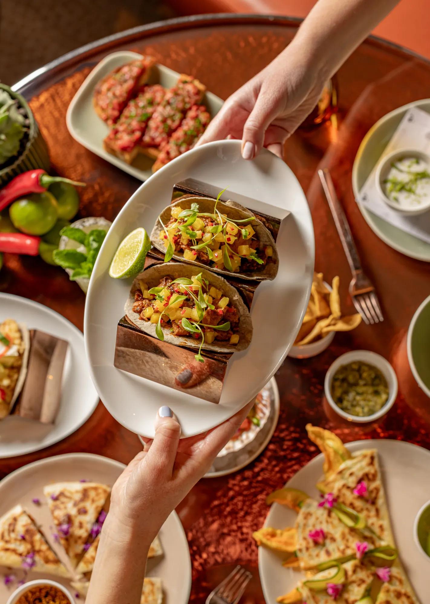 Hands present a platter of tacos above a table filled with shared Mexican dishes, fries and colourful garnishes.