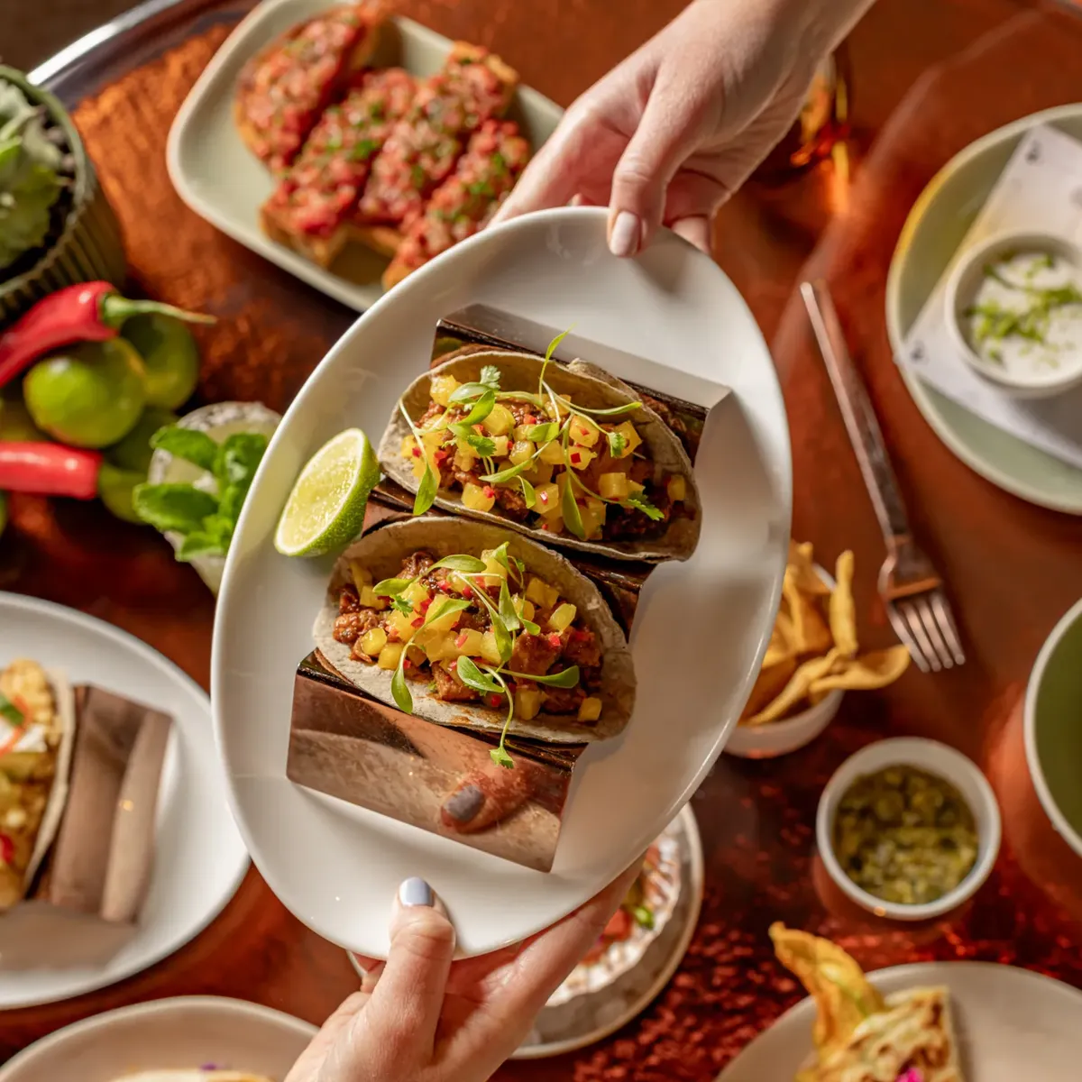 Hands present a platter of tacos above a table filled with shared Mexican dishes, fries and colourful garnishes.