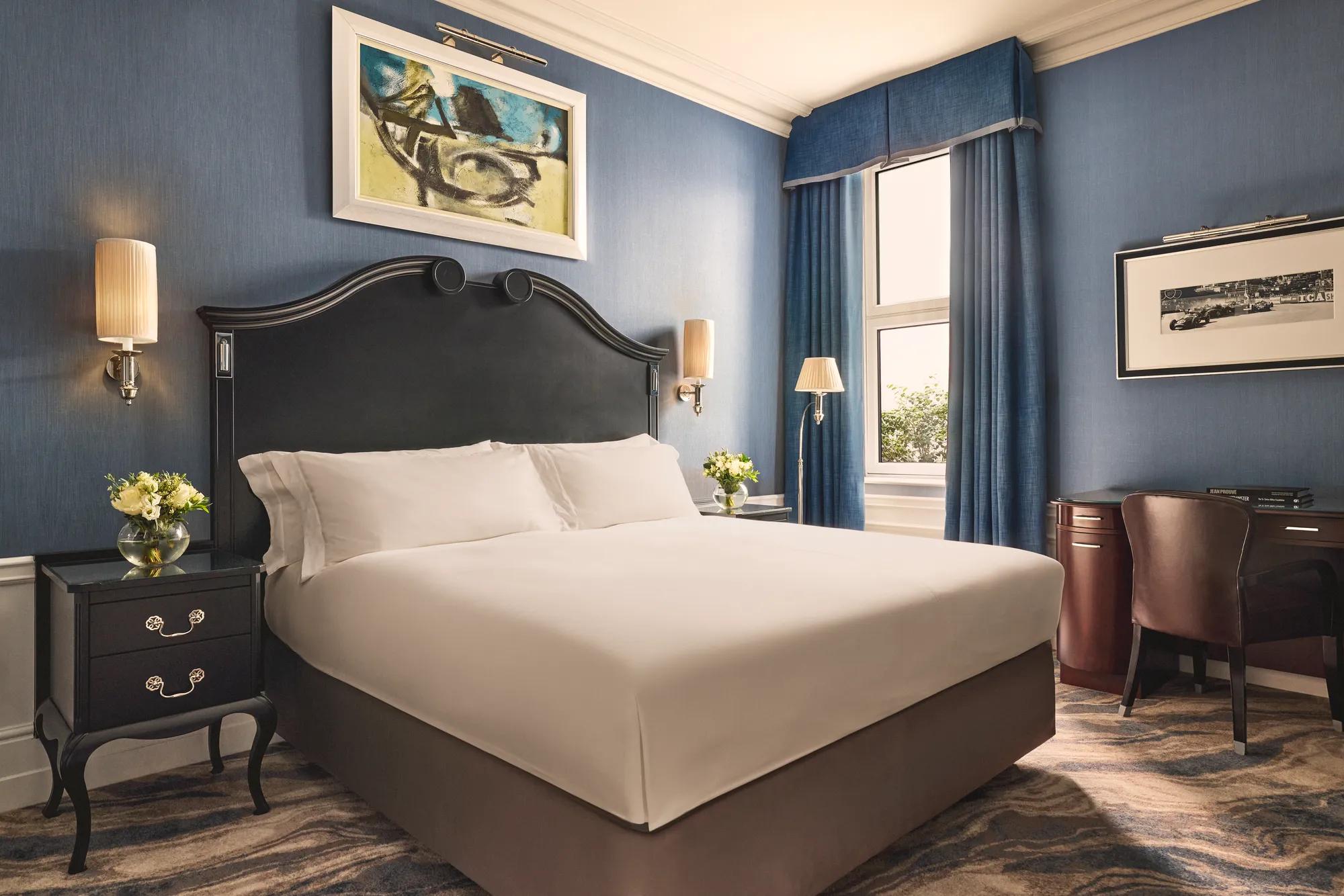 Elegant bedroom with a dark wood bed frame, crisp white bedding, blue walls and curtains, and matching bedside tables with flowers.