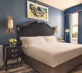 Elegant bedroom with a dark wood bed frame, crisp white bedding, blue walls and curtains, and matching bedside tables with flowers.