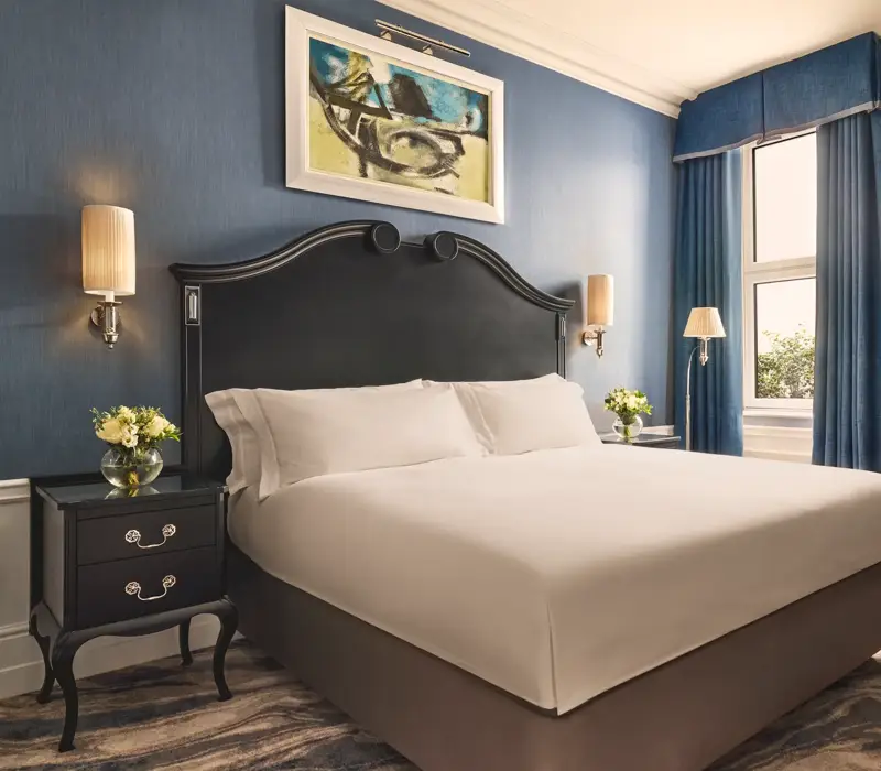 Elegant bedroom with a dark wood bed frame, crisp white bedding, blue walls and curtains, and matching bedside tables with flowers.
