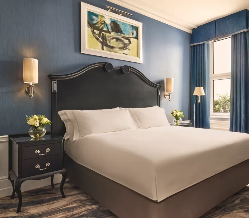 Elegant bedroom with a dark wood bed frame, crisp white bedding, blue walls and curtains, and matching bedside tables with flowers.