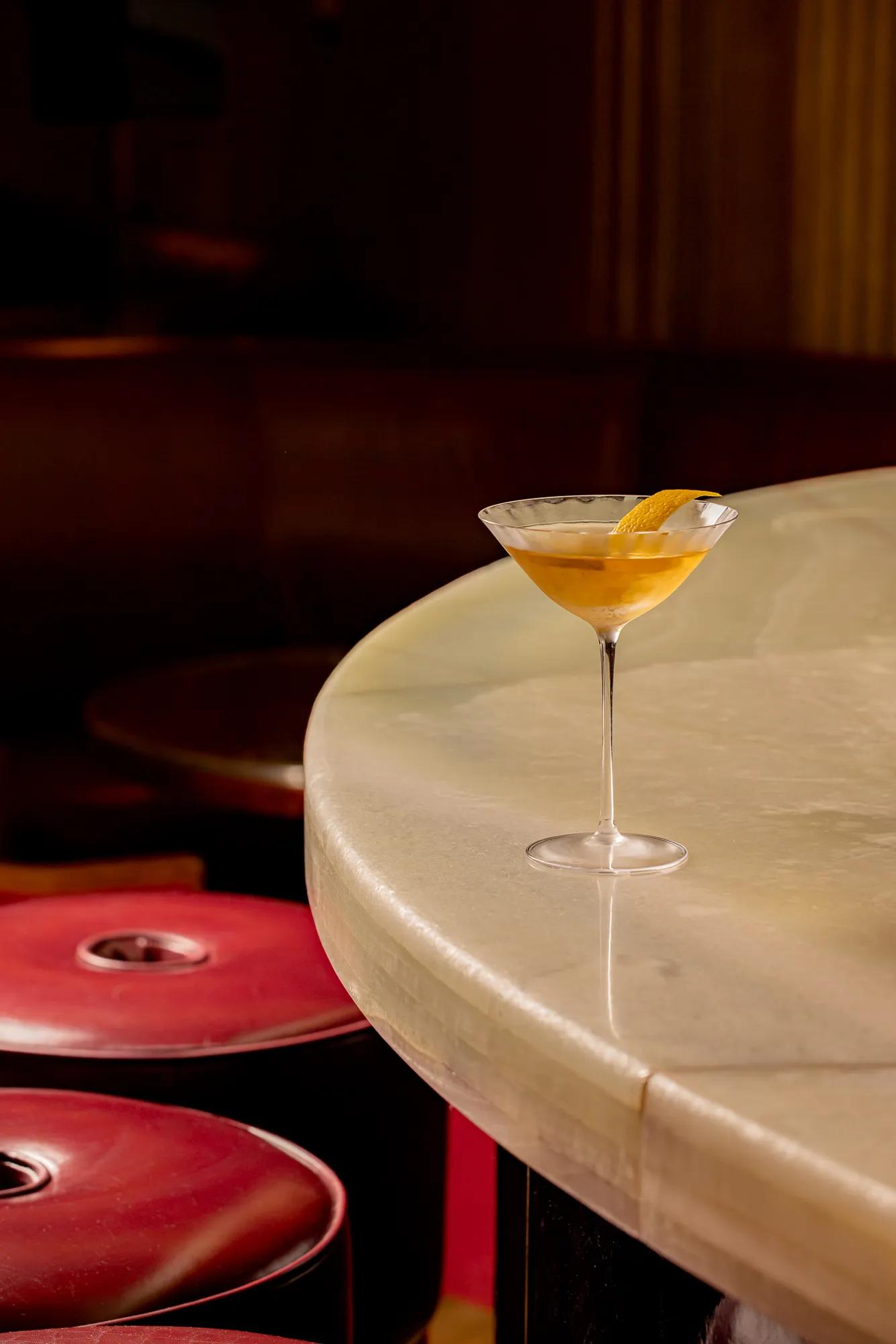 Martini glass with a golden cocktail and an orange twist garnish set on a curved marble bar counter, with red leather bar stools softly lit in the background.