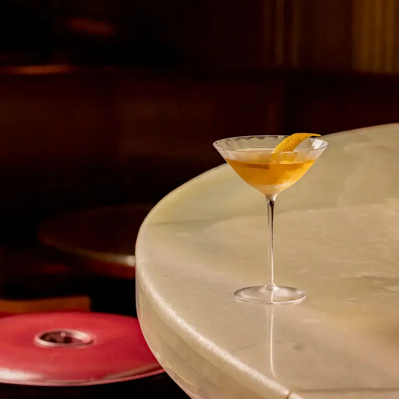 Martini glass with a golden cocktail and an orange twist garnish set on a curved marble bar counter, with red leather bar stools softly lit in the background.