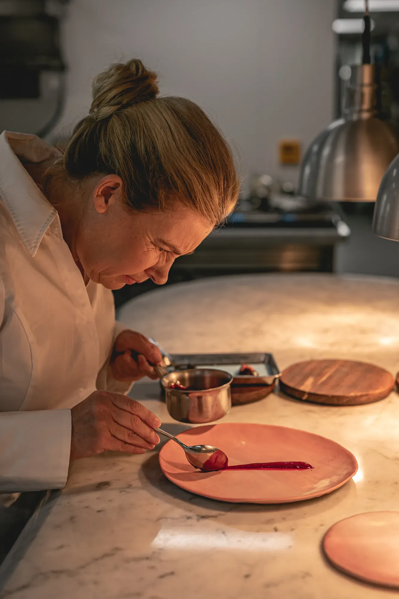 Chef plating dessert on pink plate, carefully spooning sauce with precision, lit by warm kitchen lighting and surrounded by utensils.