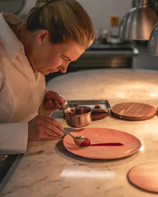 Chef plating dessert on pink plate, carefully spooning sauce with precision, lit by warm kitchen lighting and surrounded by utensils.