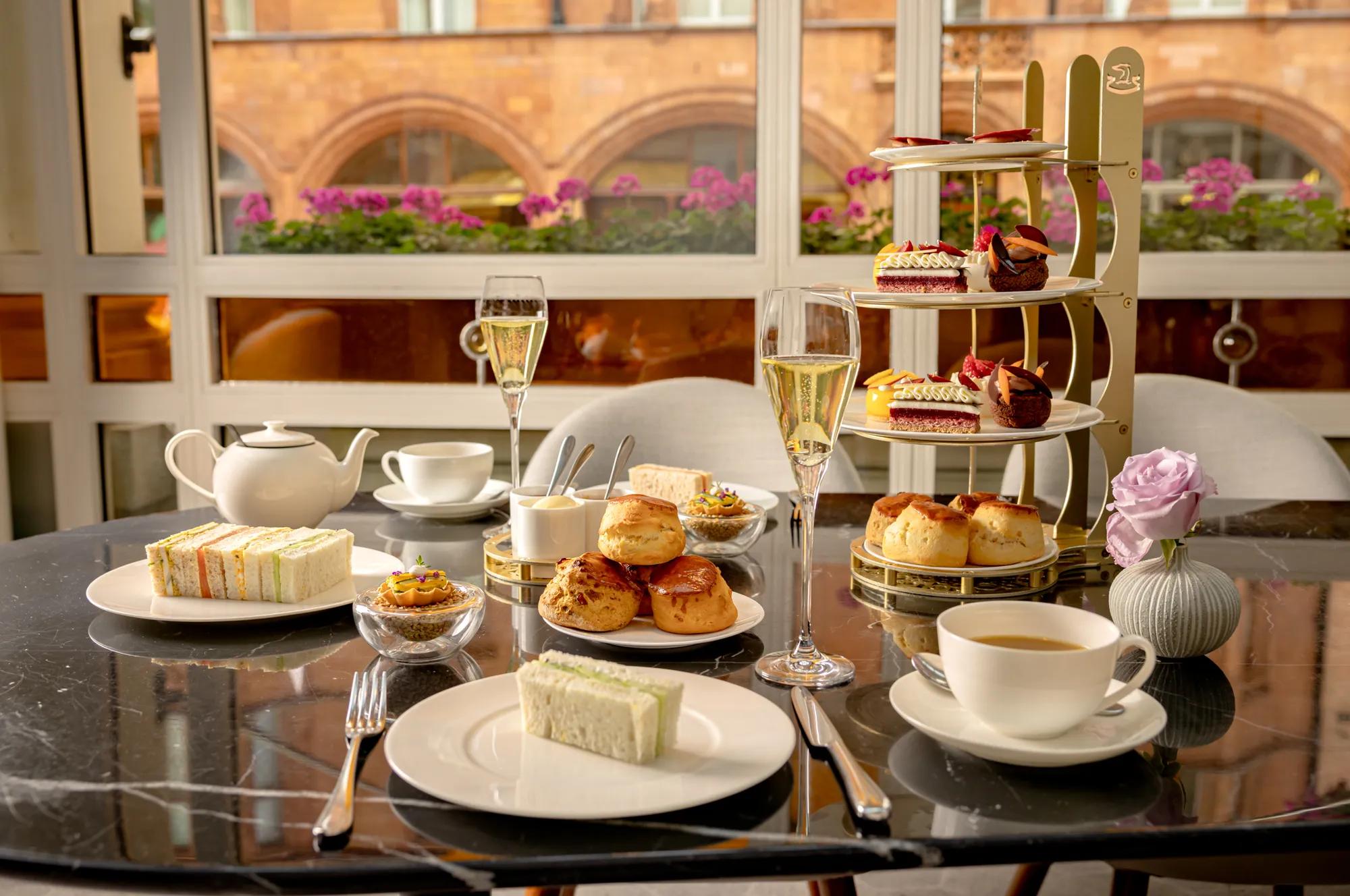 Afternoon tea with sandwiches, scones, pastries, tea and champagne served on a table beside large windows.