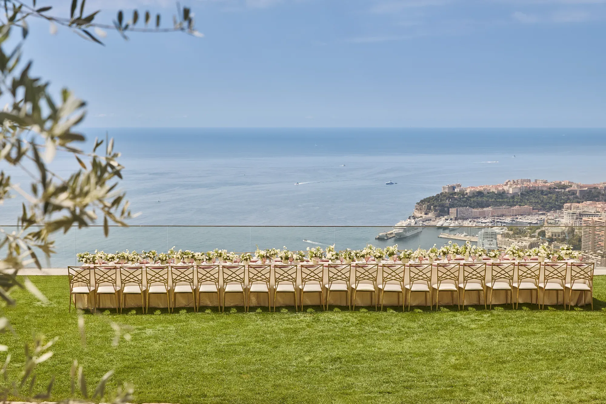 Une longue table de réception en extérieur décorée de fleurs sur une pelouse, avec vue sur la mer et le littoral de Monaco.