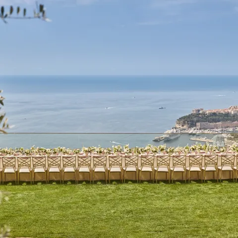 Une longue table de réception en extérieur décorée de fleurs sur une pelouse, avec vue sur la mer et le littoral de Monaco.