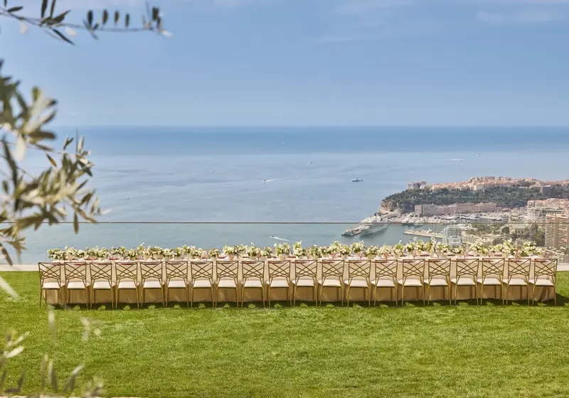 Une longue table de réception en extérieur décorée de fleurs sur une pelouse, avec vue sur la mer et le littoral de Monaco.