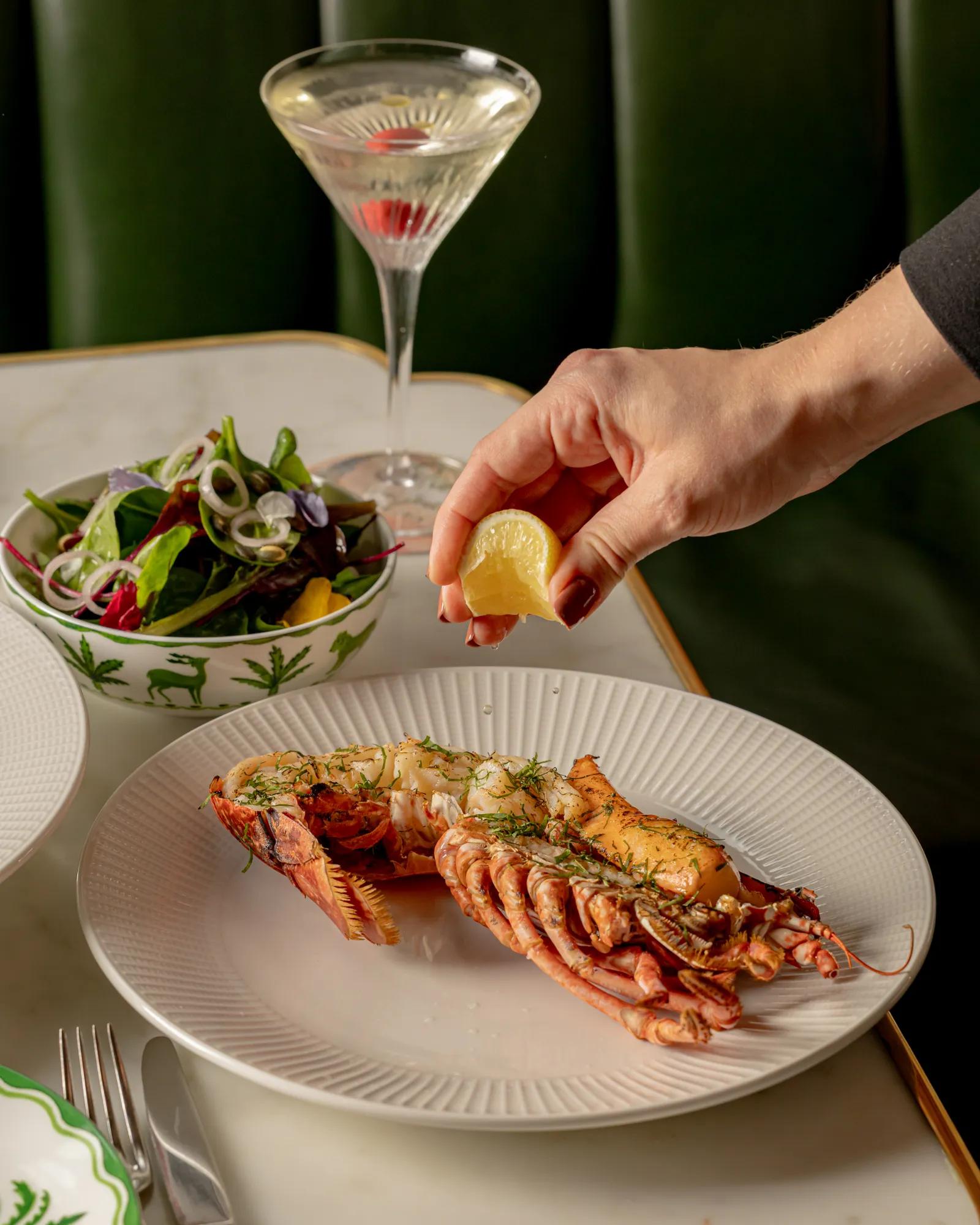 Grilled lobster on a plate with a hand squeezing a lemon wedge above it.