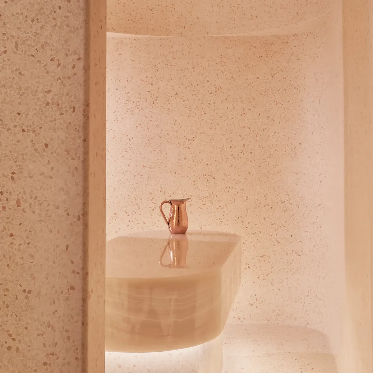 Minimalist spa steam room with smooth, curved beige stone walls and a central stone table topped with a copper water jug.