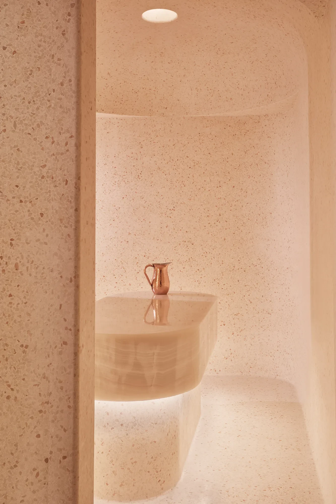 Minimalist spa steam room with smooth, curved beige stone walls and a central stone table topped with a copper water jug.