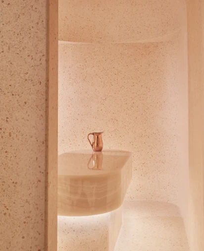Minimalist spa steam room with smooth, curved beige stone walls and a central stone table topped with a copper water jug.
