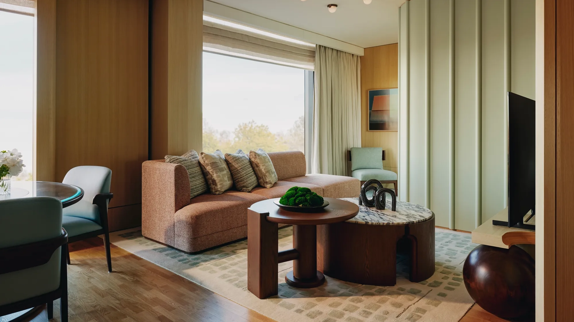 Modern suite with peach sofa, sculptural tables, and large windows overlooking trees, featuring soft lighting and warm wood tones.