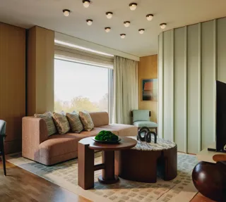 Modern suite with peach sofa, sculptural tables, and large windows overlooking trees, featuring soft lighting and warm wood tones.