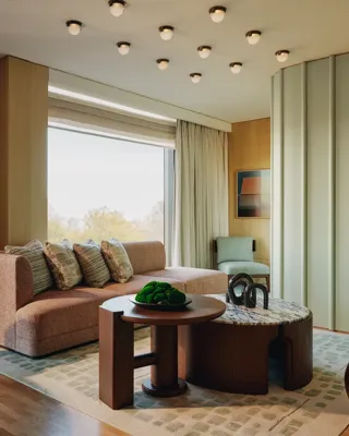 Modern suite with peach sofa, sculptural tables, and large windows overlooking trees, featuring soft lighting and warm wood tones.
