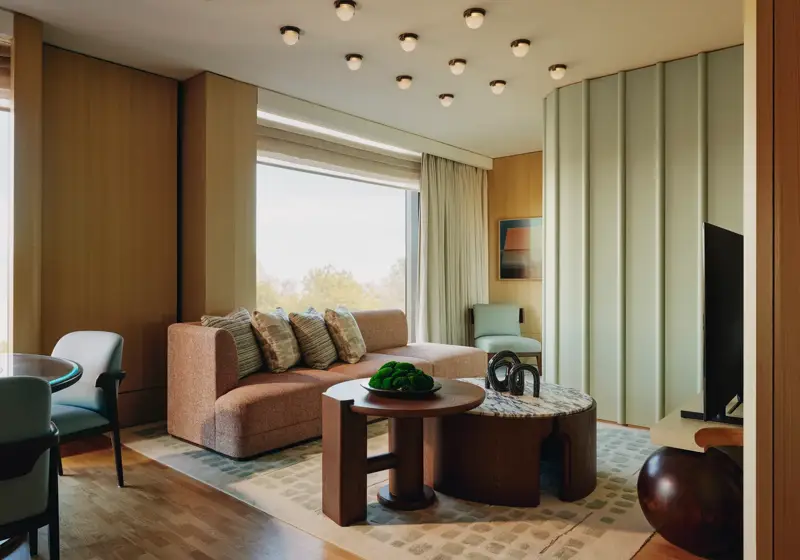 Modern suite with peach sofa, sculptural tables, and large windows overlooking trees, featuring soft lighting and warm wood tones.