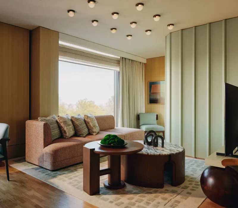 Modern suite with peach sofa, sculptural tables, and large windows overlooking trees, featuring soft lighting and warm wood tones.