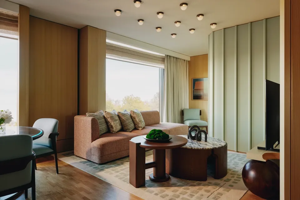 Modern suite with peach sofa, sculptural tables, and large windows overlooking trees, featuring soft lighting and warm wood tones.