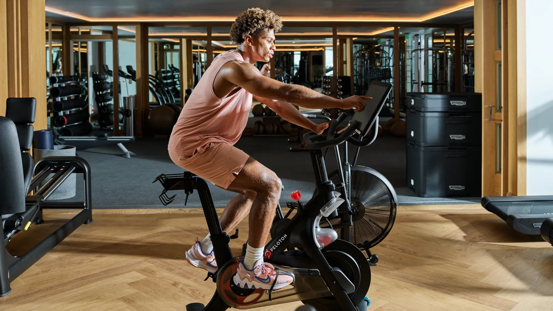 Man cycling on stationary bike in Maybourne Beverly Hills fitness center with weights and gym equipment.