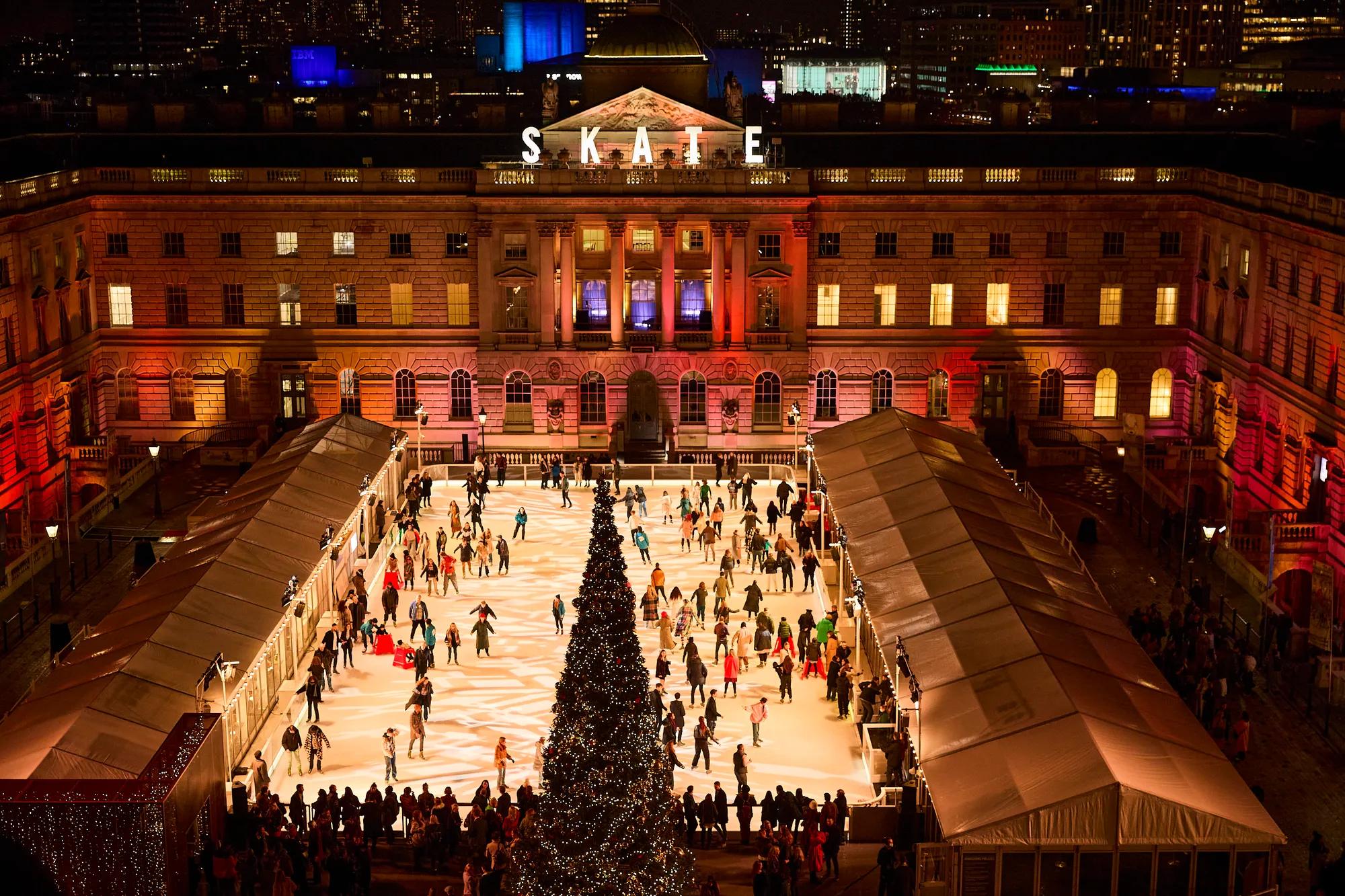 Outdoor ice rink in a grand courtyard with a central Christmas tree and skaters at night.