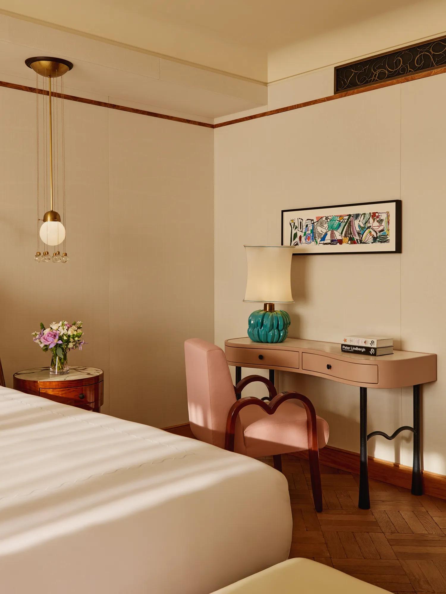 Chic bedroom corner with a blush-pink writing desk and matching chair, turquoise lamp, and framed artwork above, softly lit by natural light beside a round bedside table with flowers.
