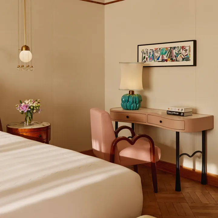 Chic bedroom corner with a blush-pink writing desk and matching chair, turquoise lamp, and framed artwork above, softly lit by natural light beside a round bedside table with flowers.