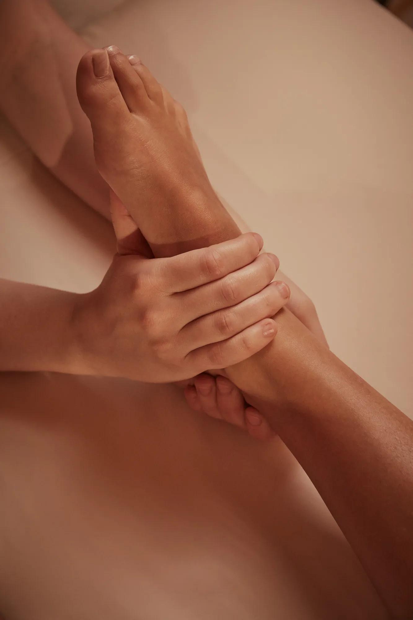 Spa therapist performing foot and ankle massage on guest in warm-toned treatment room.
