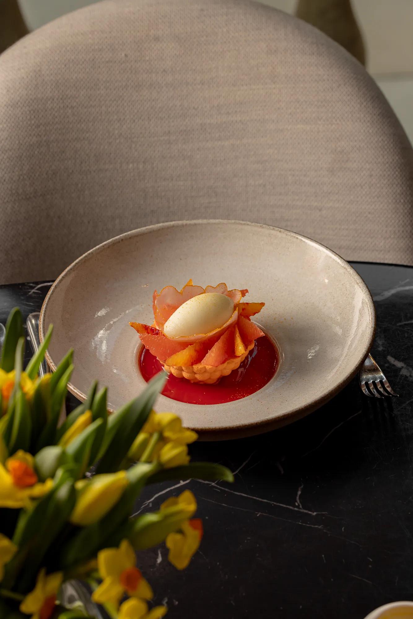 A delicate dessert of citrus segments and sorbet arranged in a flower shape, served in a shallow bowl on a dark marble table.