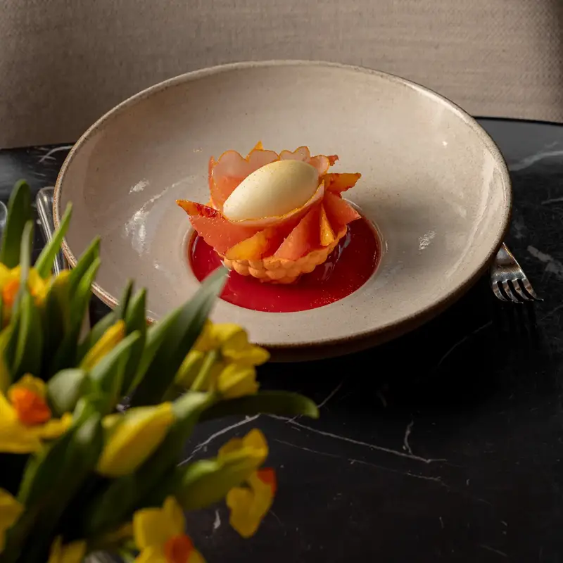 A delicate dessert of citrus segments and sorbet arranged in a flower shape, served in a shallow bowl on a dark marble table.