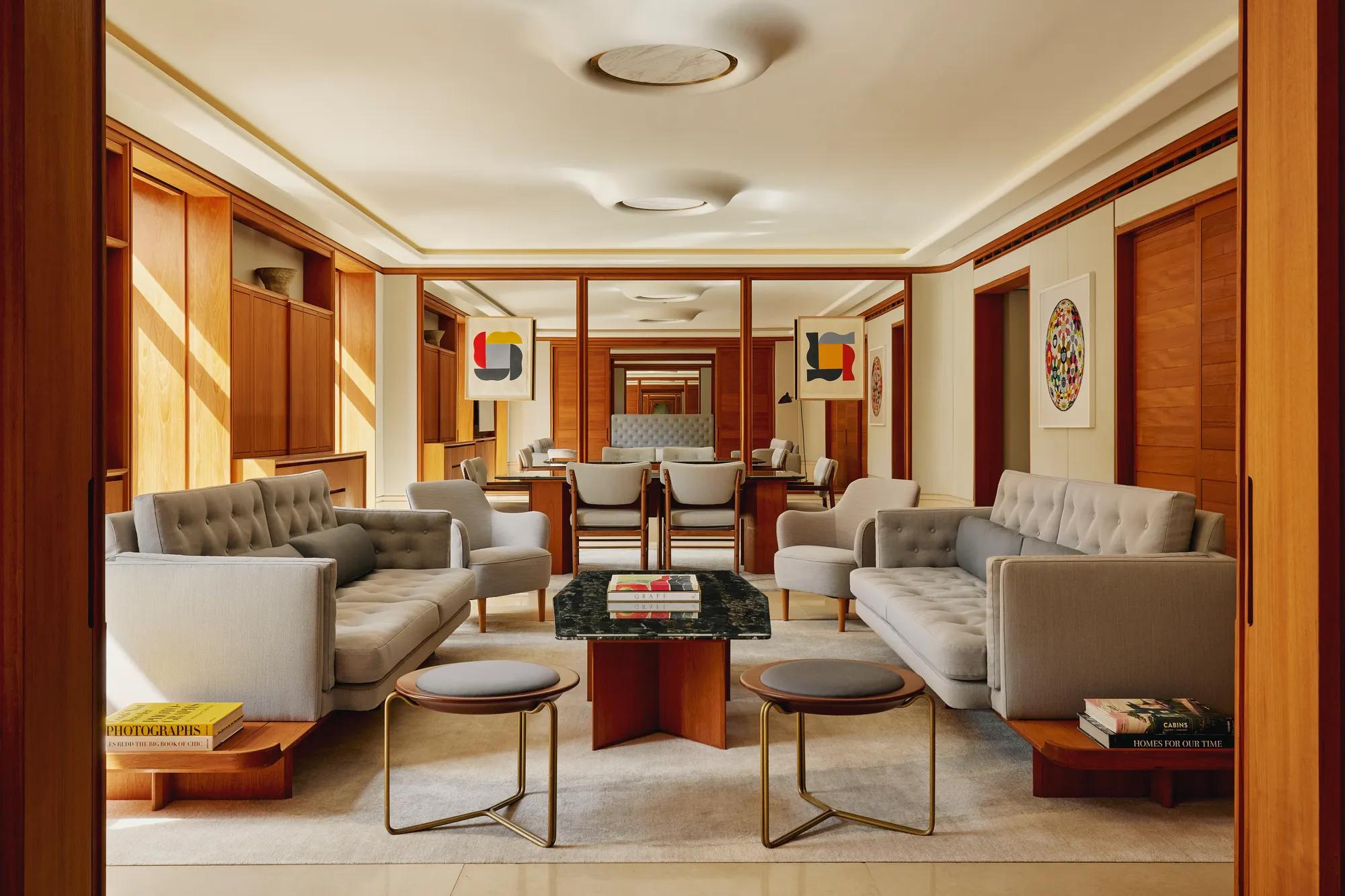 Contemporary living room in The Apartment with pale grey tufted sofas, armchairs, and a marble-topped coffee table, decorated with abstract art and wood paneling.