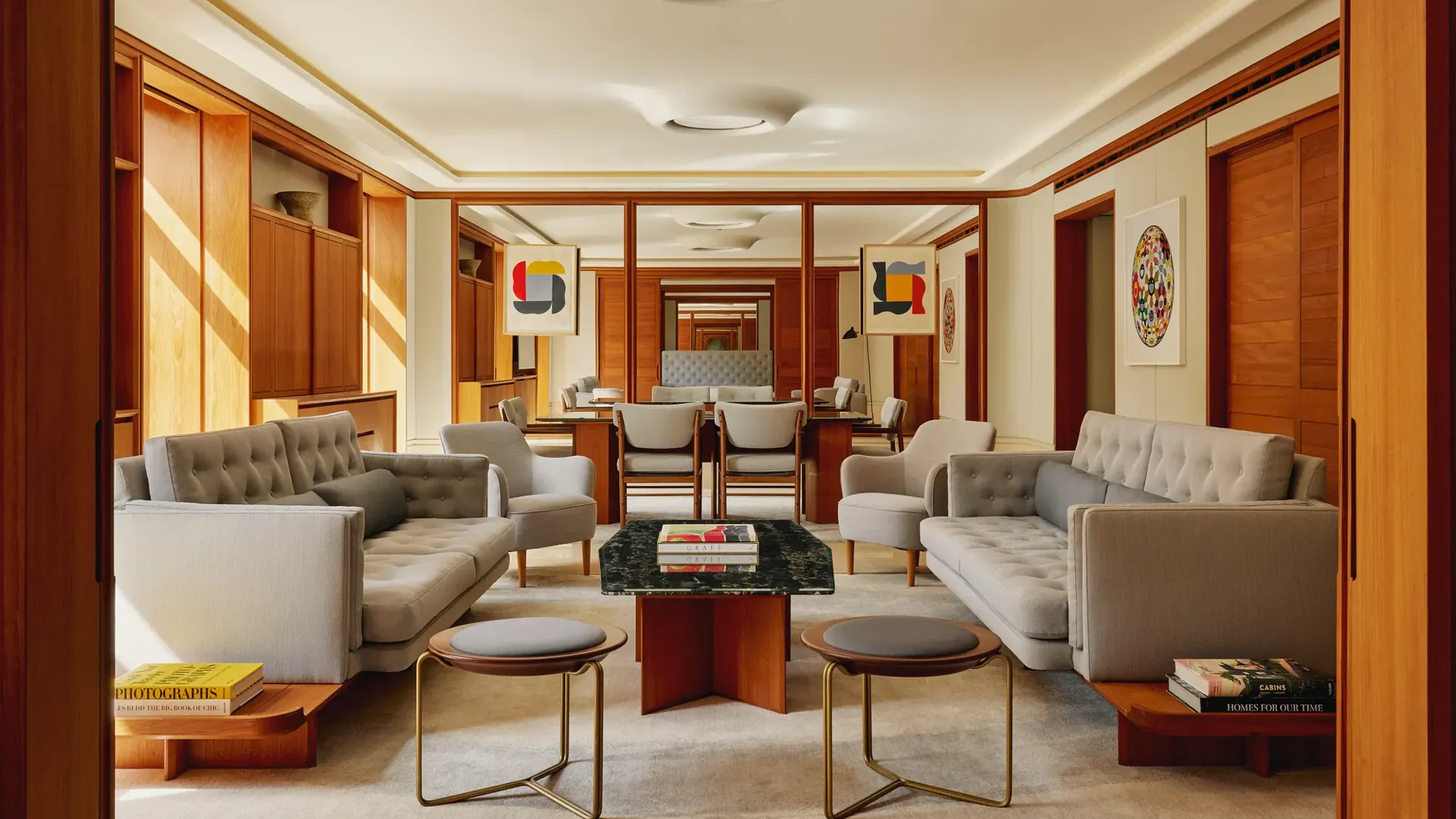 Contemporary living room in The Apartment with pale grey tufted sofas, armchairs, and a marble-topped coffee table, decorated with abstract art and wood paneling.