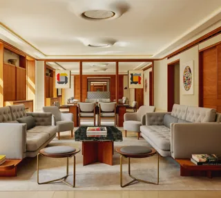 Contemporary living room in The Apartment with pale grey tufted sofas, armchairs, and a marble-topped coffee table, decorated with abstract art and wood paneling.