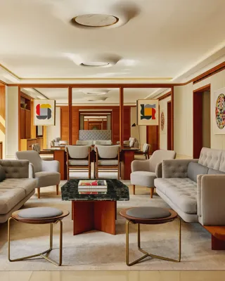Contemporary living room in The Apartment with pale grey tufted sofas, armchairs, and a marble-topped coffee table, decorated with abstract art and wood paneling.