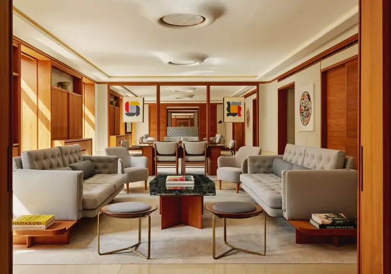 Contemporary living room in The Apartment with pale grey tufted sofas, armchairs, and a marble-topped coffee table, decorated with abstract art and wood paneling.