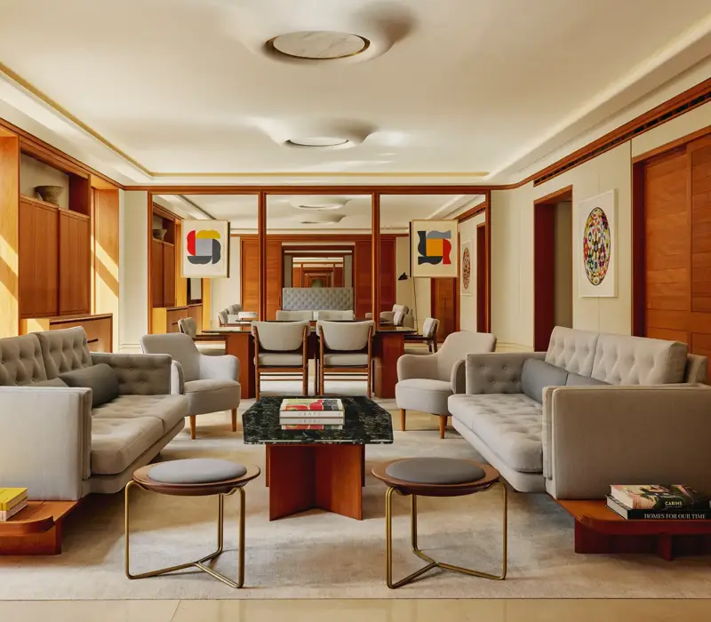 Contemporary living room in The Apartment with pale grey tufted sofas, armchairs, and a marble-topped coffee table, decorated with abstract art and wood paneling.
